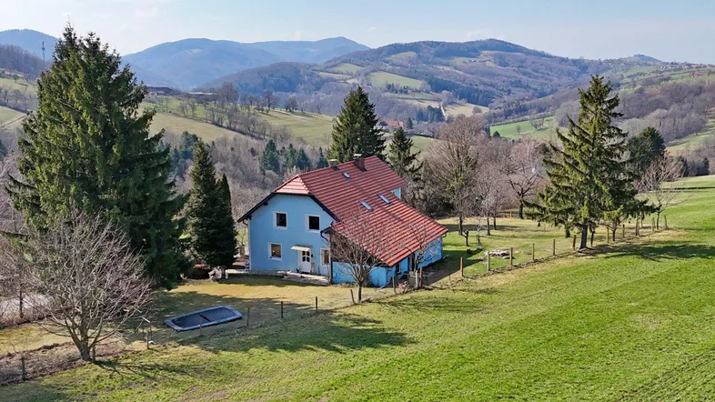 Blaues Haus mit rotem Dach in ländlicher Umgebung, umgeben von grünen Hügeln und Bäumen.