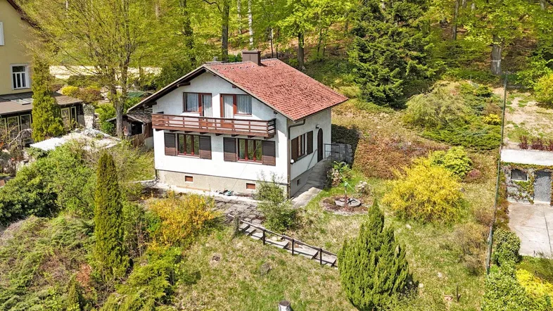 Freistehendes Einfamilienhaus mit rotem Ziegeldach und Balkon, umgeben von viel Grün und Bäumen.