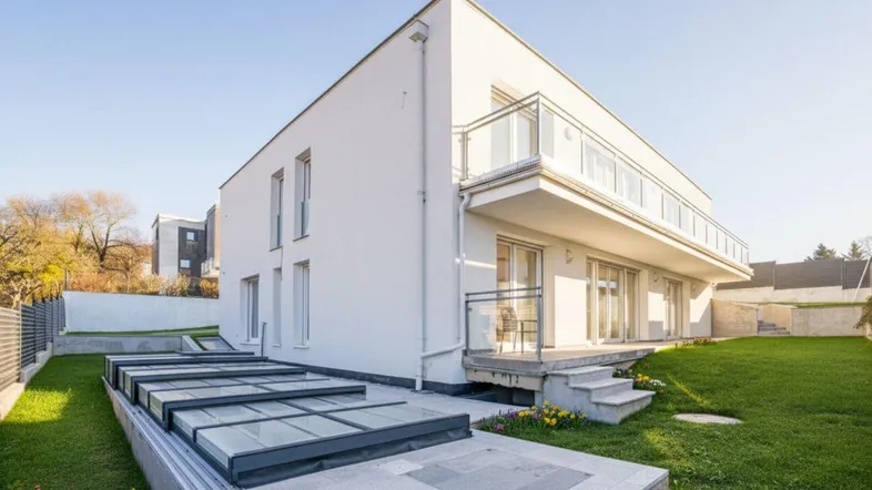 Modernes weißes Haus mit Balkon, Rasenfläche und überdachtem Poolbereich im Garten unter blauem Himmel.