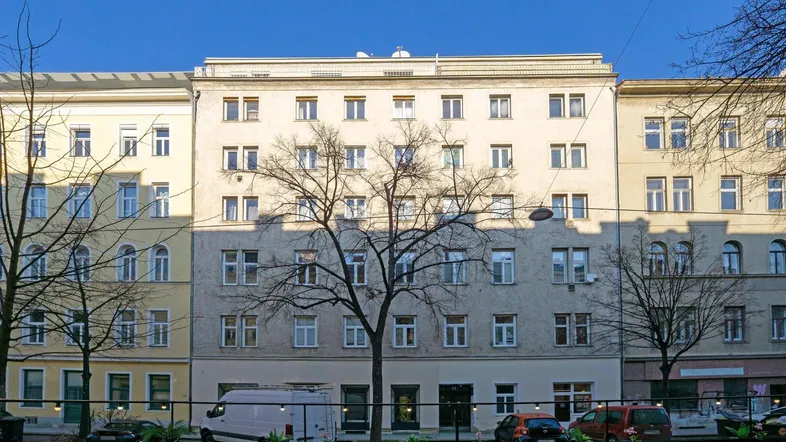Fassade eines Mehrfamilienhauses mit zahlreichen Fenstern und Bäumen davor in städtischer Umgebung.