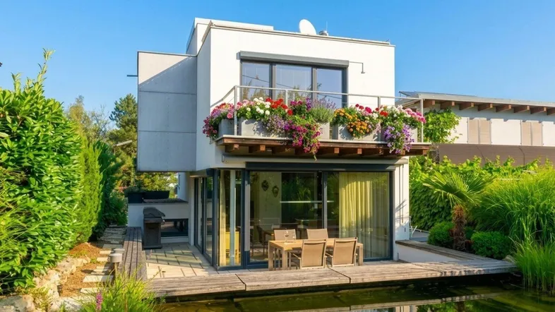 Moderne Hausfassade mit Teich, Terrasse und blumengeschmücktem Balkon unter blauem Himmel.