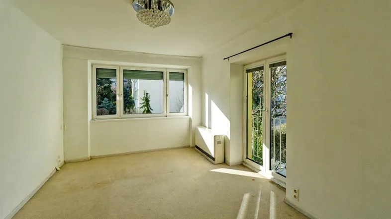 Heller Raum mit Teppichboden, großen Fenstern, Heizkörper und Zugang zu einem französischen Balkon mit Grünblick.