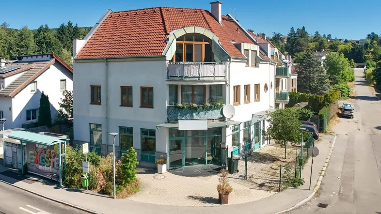 Mehrstöckiges Gebäude mit rotem Ziegeldach, großen Fenstern und einem kleinen Balkon an einer Straßenecke.