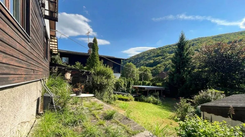 Seitlicher Blick auf das Haus mit Holzfassade und einem naturbelassenen Gartenbereich am Hang.