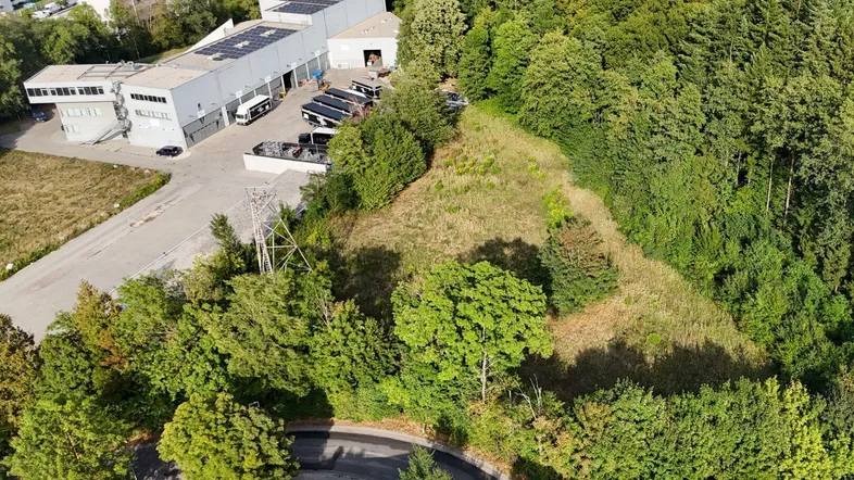 Luftaufnahme eines Grundstücks umgeben von Wald und Gewerbegebäuden, mit einer Zufahrtsstraße im Vordergrund.