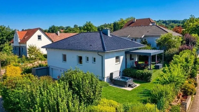 Luftaufnahme des Hauses mit gepflegtem Garten, überdachter Terrasse und Whirlpool unter blauem Himmel.