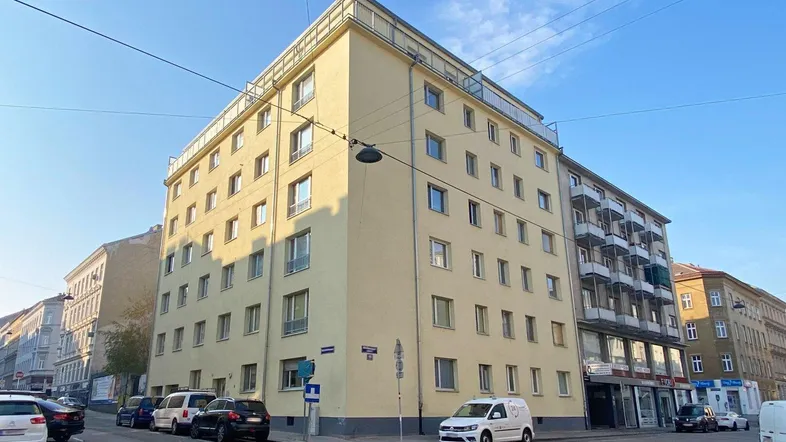 Mehrstöckiges Wohngebäude mit heller Fassade an einer Straßenecke, umgeben von parkenden Autos und städtischer Architektur.