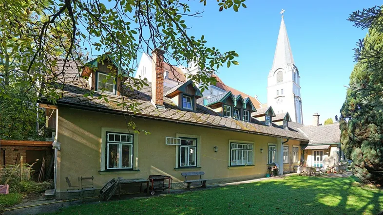 Gelbes Haus mit Satteldach und Gauben, umgeben von einem grünen Garten, mit einer Kirche im Hintergrund.