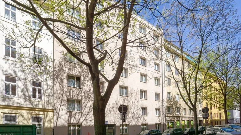 Mehrstöckiges Wohngebäude mit heller Fassade und vielen Fenstern, umgeben von Bäumen an einer Stadtstraße.