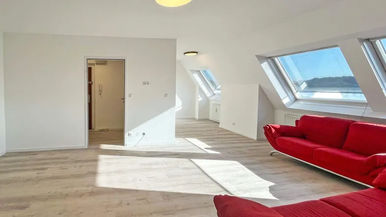 Helles Dachgeschosszimmer mit Schrägfenstern, Laminatboden und rotem Sofa, durchflutet von Sonnenlicht.