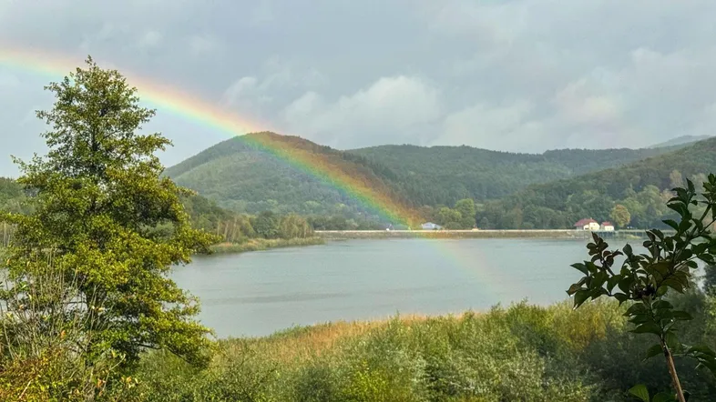 Malerische Landschaft mit See, bewaldeten Hügeln und einem Regenbogen am Himmel.