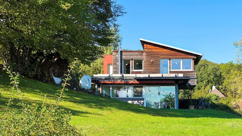 Modernes Haus mit Holzfassade und großen Glasfronten, eingebettet in eine grüne Hügellandschaft.