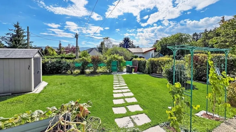 Grüner Garten mit Rasenfläche, Steinweg und Gartenhaus unter blauem Himmel.