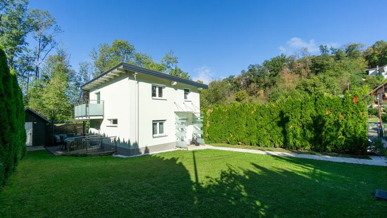 Weißes Einfamilienhaus mit gepflegtem Rasen und hohen Hecken unter blauem Himmel.