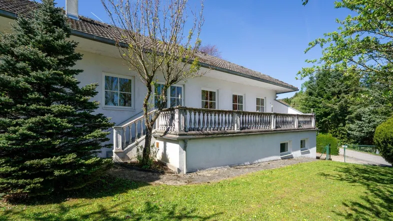 Traditionelles weißes Haus mit Balkon und Balustrade, umgeben von einem gepflegten grünen Garten unter blauem Himmel.