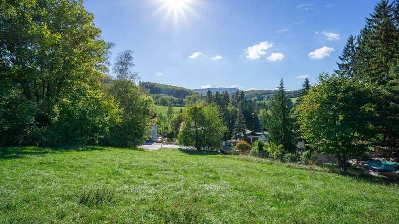 Weitläufige grüne Landschaft mit sonnigem Himmel und Blick auf bewaldete Hügel und vereinzelte Häuser.