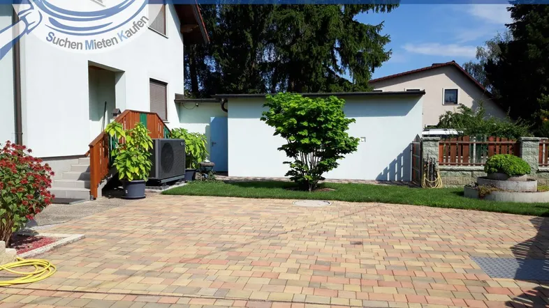 Gepflasterter Vorplatz mit Rasenfläche und Bäumen vor dem Haus an einem sonnigen Tag.