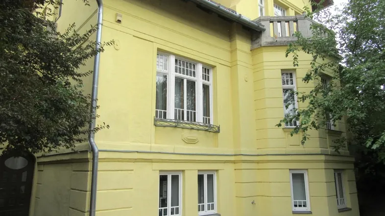 Gelbe Fassade eines Altbaus mit klassischen weißen Fenstern und einem kleinen dekorativen Balkon.