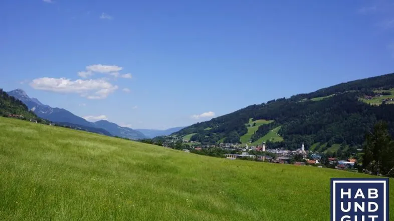 Weitläufige grüne Wiesen mit einem Dorf und Bergen im Hintergrund unter blauem Himmel.