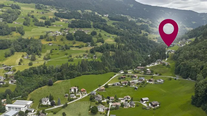 Luftaufnahme eines grünen Tals mit verstreuten Häusern und Bergen im Hintergrund, markiert mit einem Standort-Pin.