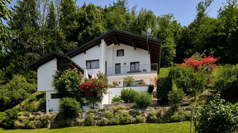Weißes Einfamilienhaus mit Satteldach, umgeben von üppigem Grün und einem gepflegten Garten.