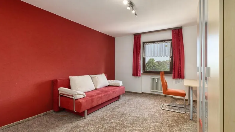 Heller Raum mit roter Akzentwand, Schlafsofa und Schreibtisch, ideal als Gästezimmer.