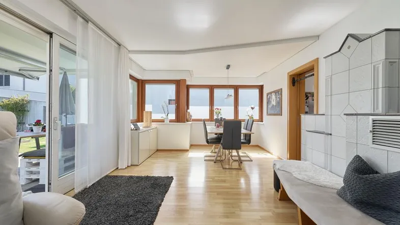 Heller Wohn- und Essbereich mit Parkettboden, Kachelofen und Zugang zur Terrasse.