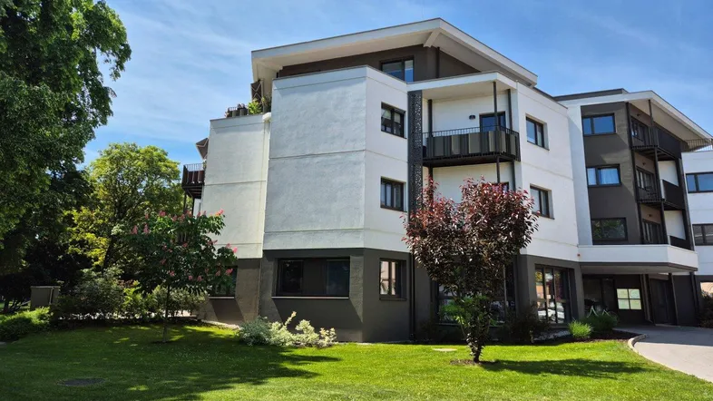 Modernes Apartmentgebäude mit weißer und grauer Fassade, umgeben von Grünflächen und Bäumen.