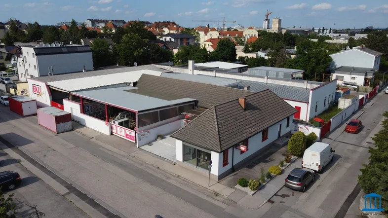 Luftaufnahme eines Gewerbeobjekts mit mehreren Gebäudeteilen und angrenzender Wohnbebauung unter blauem Himmel.