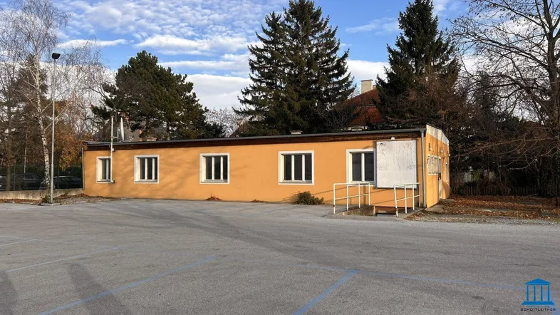 Gelbes, eingeschossiges Gebäude mit mehreren Fenstern und einem Parkplatz davor unter bewölktem Himmel.