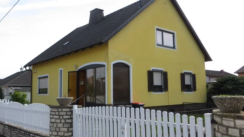 Gelbes Einfamilienhaus mit Satteldach, weißem Zaun und braunen Fensterläden.