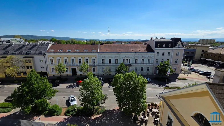 Panoramablick über eine Stadtstraße mit historischen Gebäuden und grünen Bäumen unter blauem Himmel.