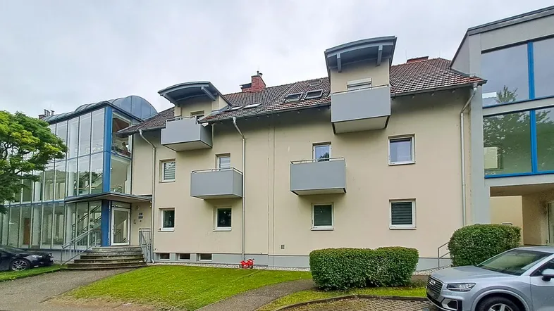 Mehrfamilienhaus mit beiger Fassade, mehreren Balkonen und einem markanten, modernen Glasanbau, umgeben von Grünflächen.