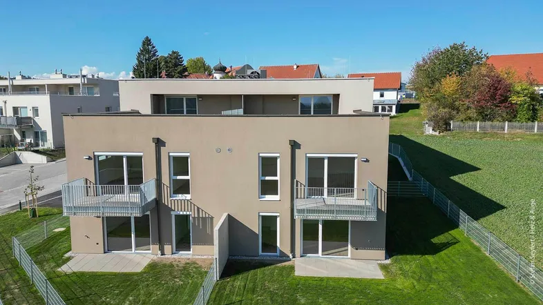 Modernes Mehrfamilienhaus mit Balkonen und Terrassen, umgeben von grünen Feldern unter blauem Himmel.