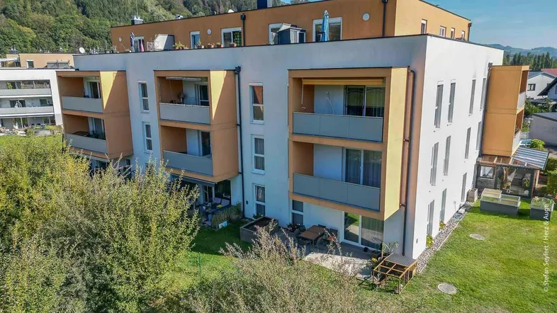 Modernes Mehrfamilienhaus mit Balkonen und Terrassen, umgeben von Grünflächen und Bergen.