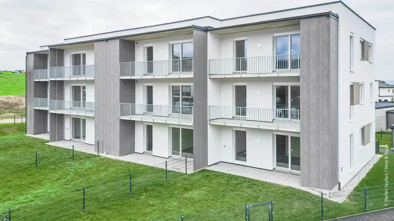 Modernes Mehrfamilienhaus mit weißer Fassade, grauen Akzenten und Balkonen in ländlicher Umgebung.