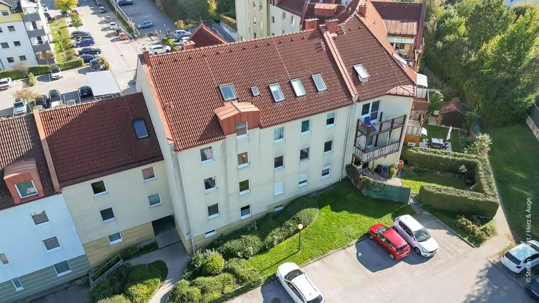 Luftaufnahme eines Mehrfamilienhauses mit rotem Ziegeldach, umgeben von Grünflächen und Parkplätzen.