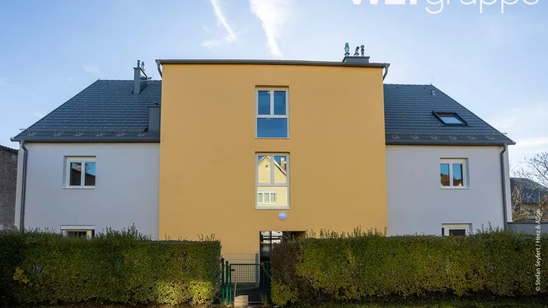 Moderne Gebäudefassade mit gelbem Mittelteil, grauen Dächern und gepflegter Hecke.
