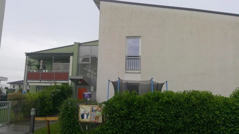 Außenansicht eines modernen Gebäudes mit Balkonen, Hecke und einem Trampolin im Gartenbereich.