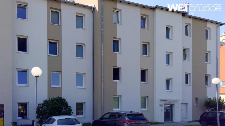 Mehrstöckiges Wohngebäude mit heller Fassade und zahlreichen Fenstern, umgeben von Parkplätzen und Grünflächen.
