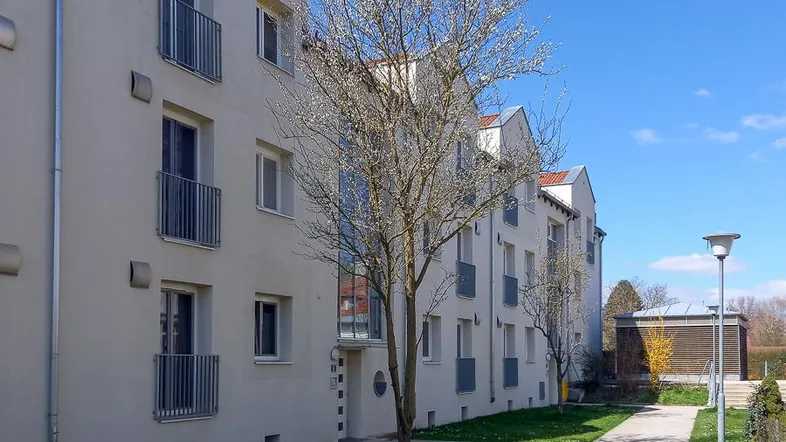 Außenansicht eines modernen Mehrfamilienhauses mit Balkonen, Grünflächen und einem Gehweg unter blauem Himmel.
