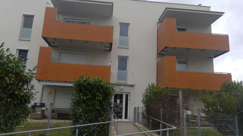 Modernes Mehrfamilienhaus mit hellgrauer Fassade, Holzbalustraden und barrierefreiem Zugang über eine Rampe.