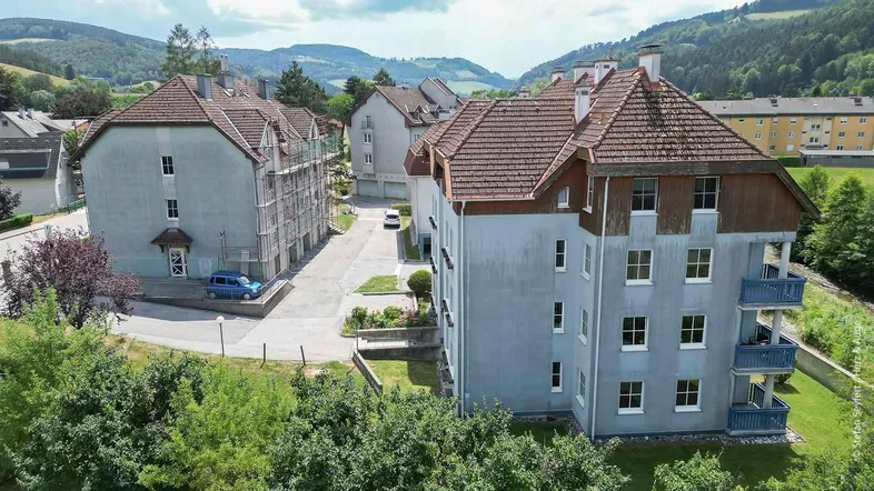 Luftaufnahme von Wohngebäuden in einer grünen, hügeligen Umgebung, eines davon in Renovierung.
