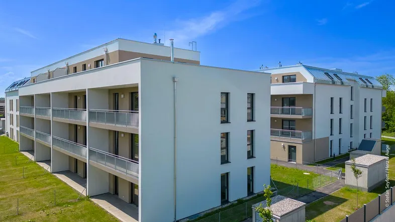 Moderne Mehrfamilienhäuser mit Balkonen und grünen Außenanlagen unter blauem Himmel.