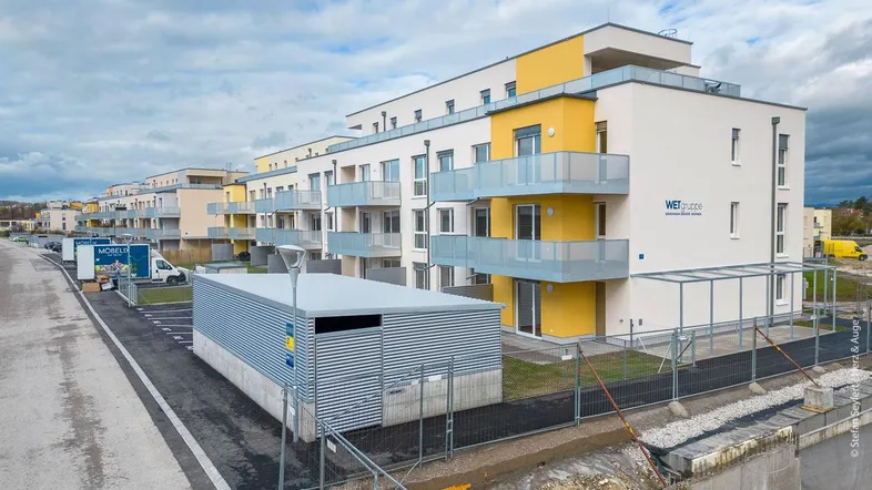 Modernes Mehrfamilienhaus mit gelben Akzenten, Balkonen und einer Neubausiedlung im Hintergrund.