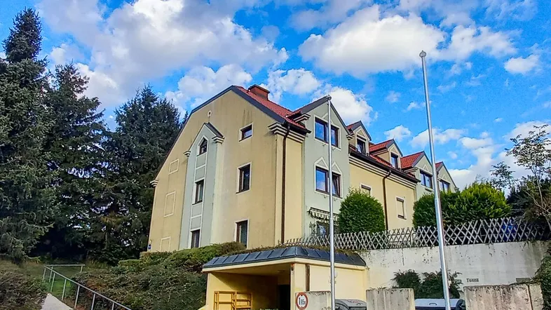 Gelbes Mehrfamilienhaus mit Satteldach und Gauben, umgeben von hohen Bäumen und gepflegten Sträuchern.