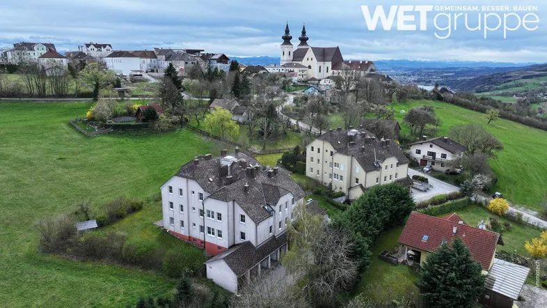 Luftaufnahme eines Wohngebäudes in ländlicher Umgebung mit Blick auf eine Kirche und grüne Hügel.