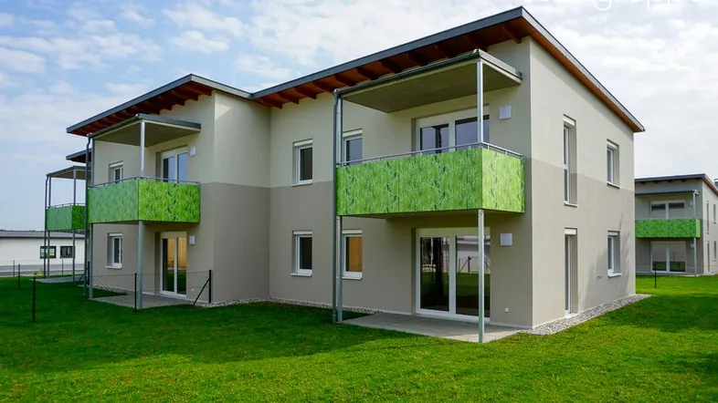 Modernes Neubau-Mehrfamilienhaus mit grünen Balkonen und gepflegter Rasenfläche unter blauem Himmel.