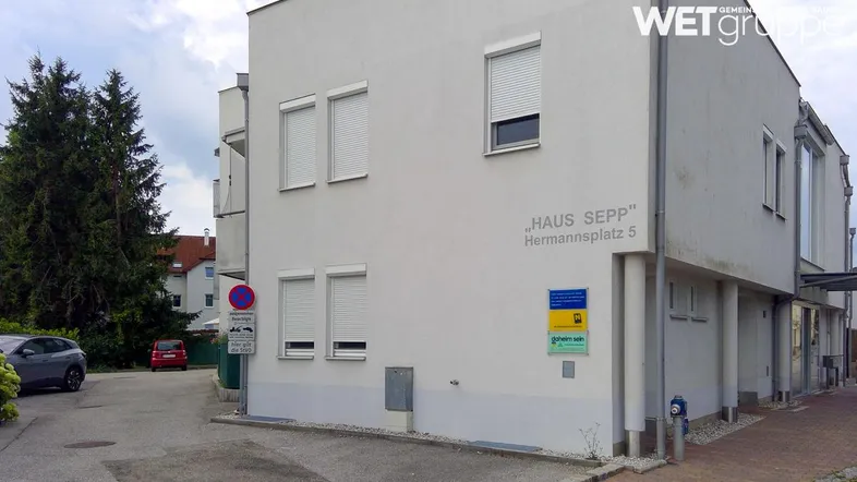Modernes Mehrfamilienhaus mit weißer Fassade, Rollläden und angrenzendem Parkplatz.