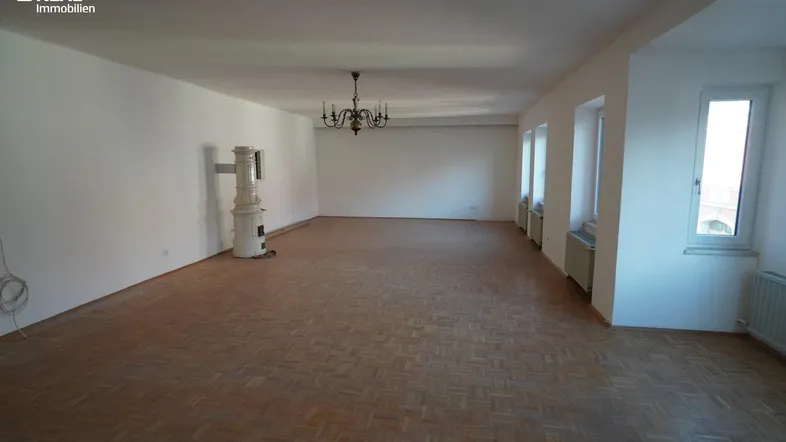 Geräumiges Wohnzimmer mit Fischgrätparkett, Kachelofen und mehreren Fenstern für viel Tageslicht.